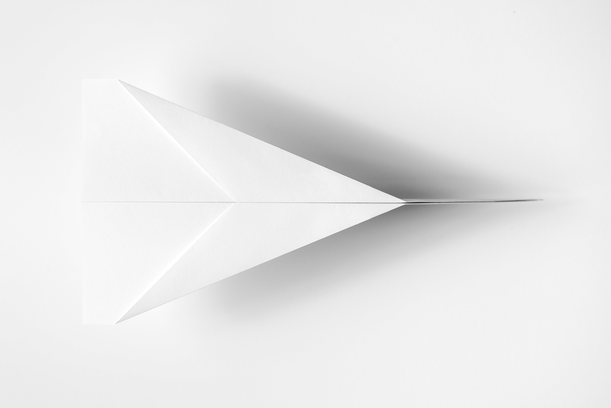 Minimalist photograph of a white paper plane viewed from above on a white background.