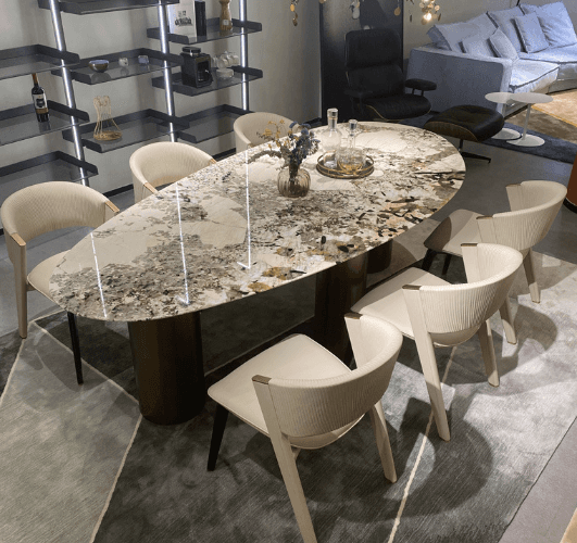 An oval marble dining table with intricate natural patterns in cream and brown tones. It is surrounded by white chairs in a modern dining room with a cozy, elegant atmosphere.