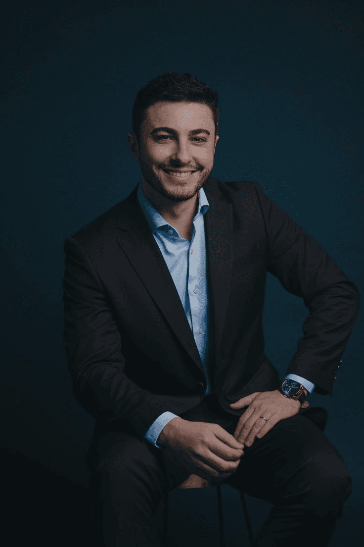 Smiling man in a dark suit and light blue shirt, seated against a dark background. His hands rest on his knee, exuding professionalism and warmth.