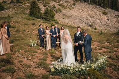 The couple and officiant stand inside a curve of white flowers as guests watch nearby.