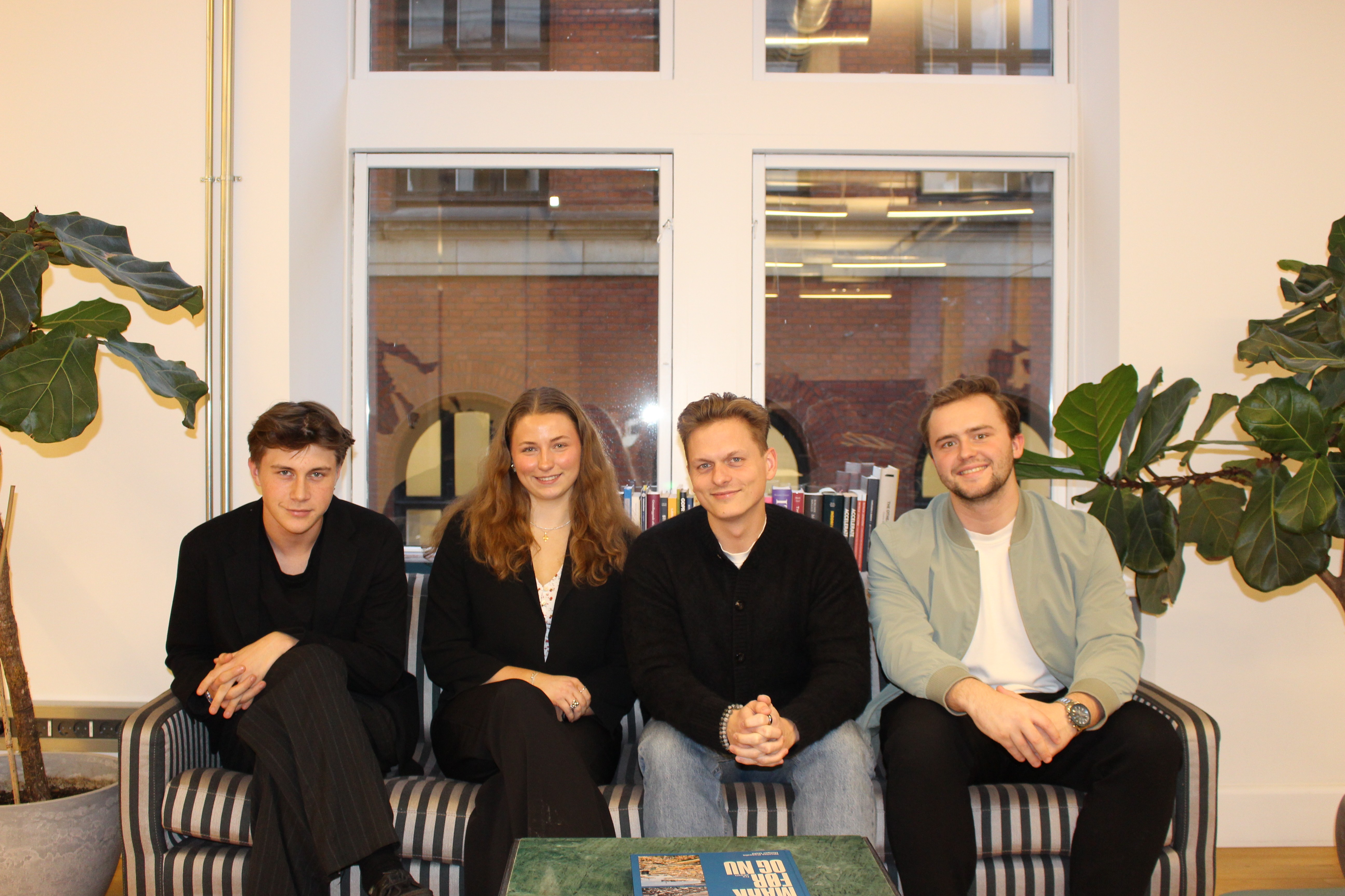 A smiling man and woman sit together on a sofa in a sunlit room, with large green potted plants in the background, embodying a warm and cheerful atmosphere.