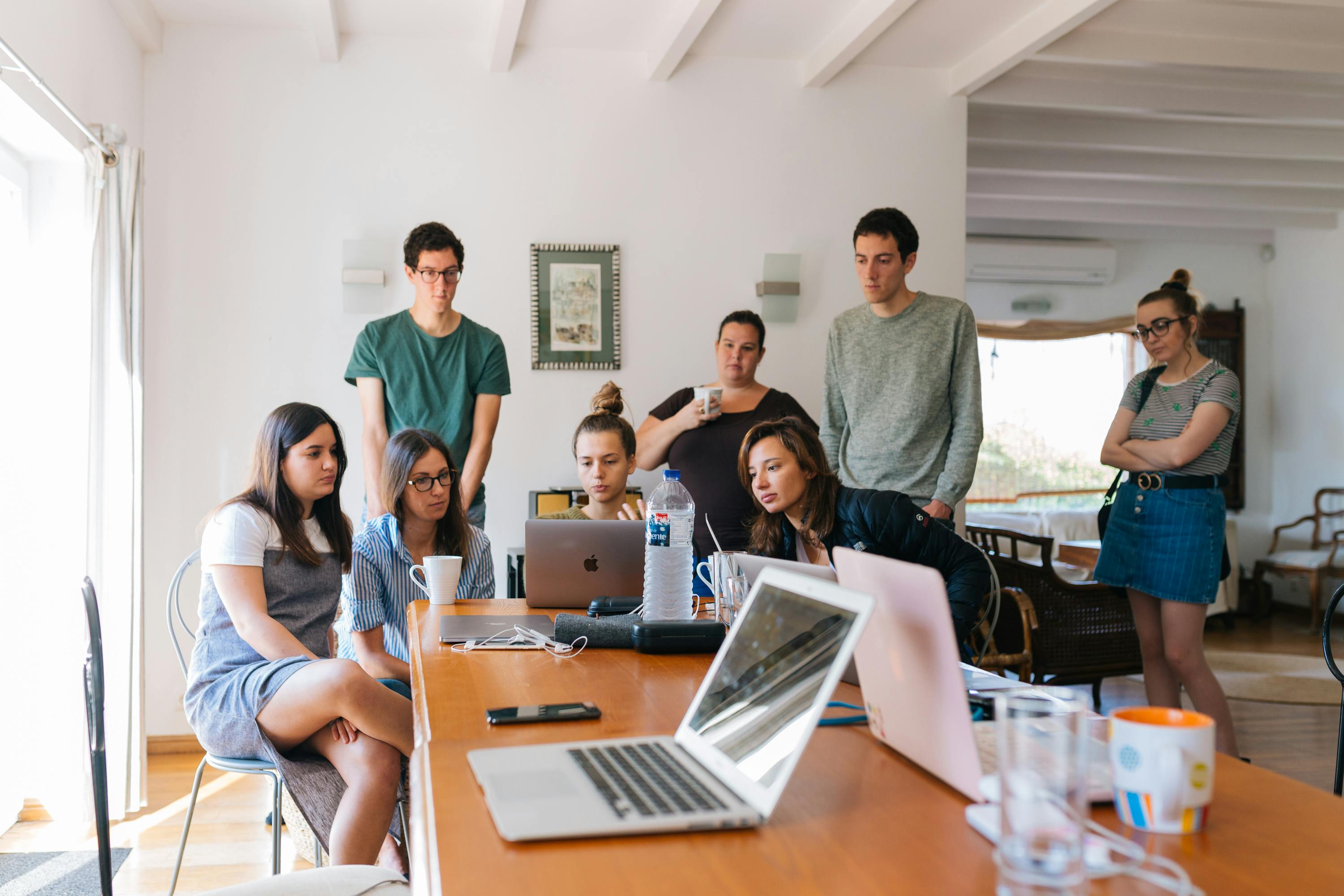 A diverse group of six people stands and sits around a table with a laptop in a bright, modern room.