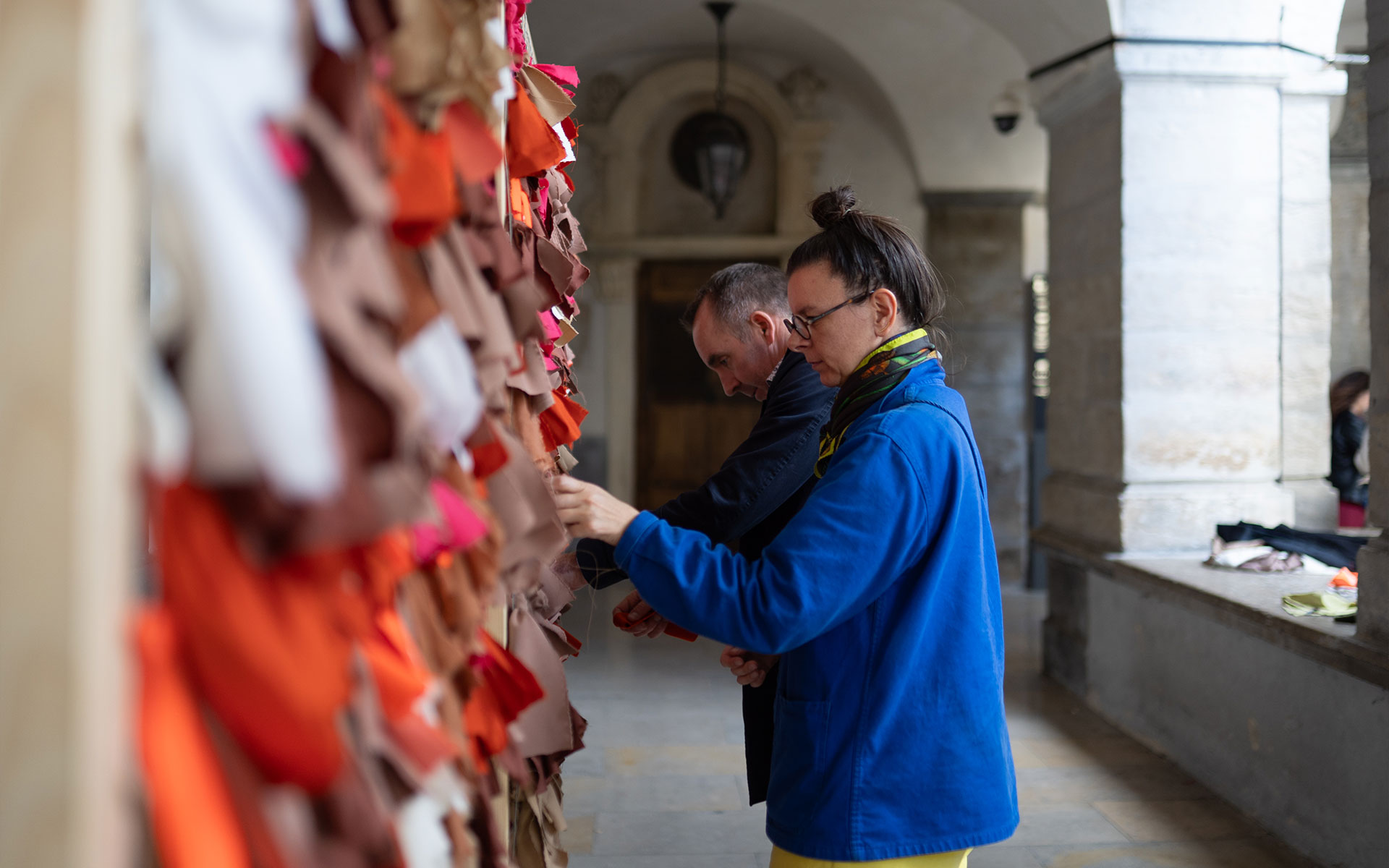 Dans un passage couvert aux colonnes en pierre, deux personnes travaillent minutieusement sur une grande installation textile composée de nombreuses bandes de tissus de diverses teintes (marron, orange, rouge, blanc). Une femme au premier plan, portant une veste bleue et des lunettes, manipule les tissus aux côtés d'un homme qui l'assiste dans cette tâche précise. La profondeur de champ met l'accent sur leur action, tandis que l'arrière-plan révèle une architecture classique et lumineuse.