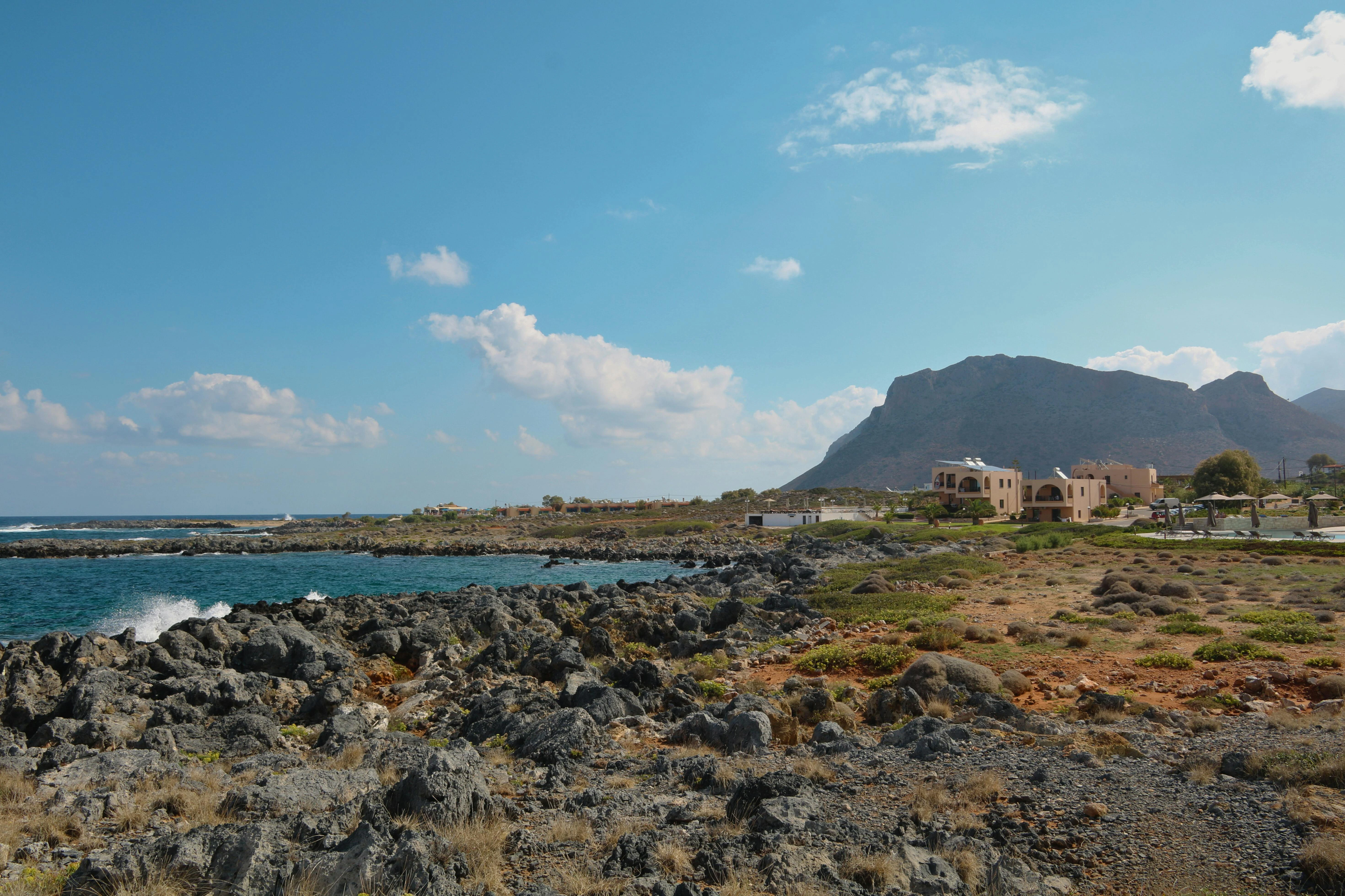 A rocky coastline with turquoise water under a blue sky, featuring a small village and a large mountain in the background.