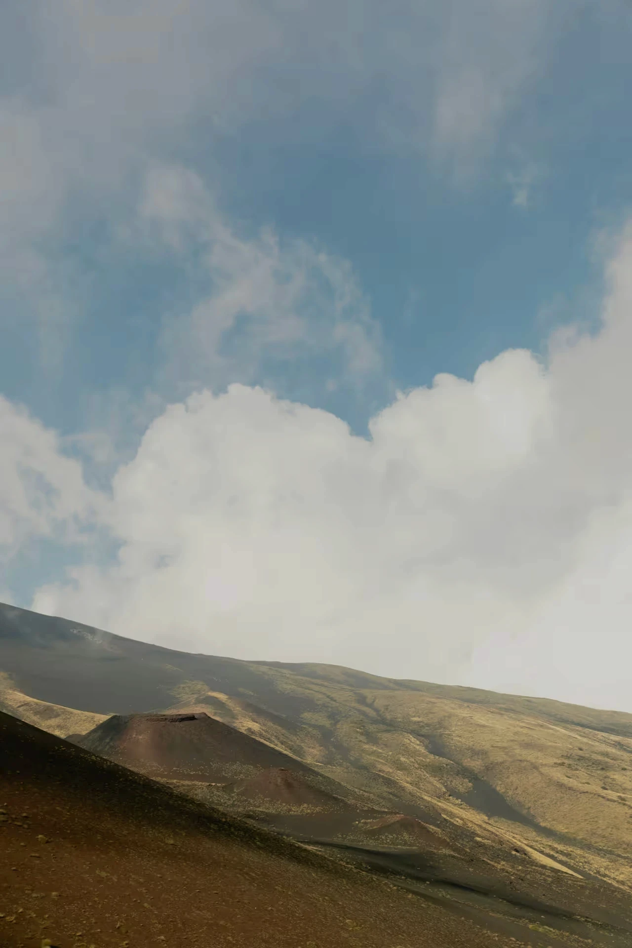 Barren volcanic hillside with a small crater under a partly cloudy blue sky.