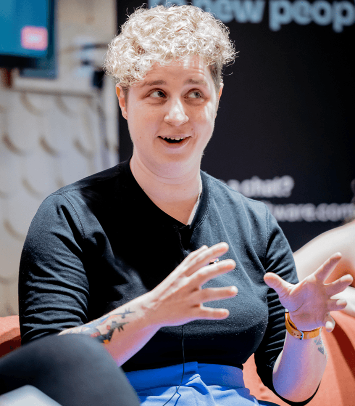 A seated person speaking on a panel or at a discussion event, gesturing with both hands while talking. They are wearing a black long‑sleeve top and blue trousers, with a visible tattoo on one forearm and a watch on the wrist. The background shows part of an indoor venue with event branding and soft lighting, suggesting a professional or community talk setting.