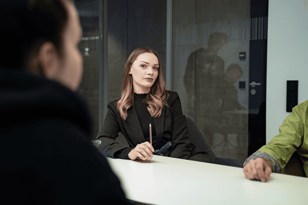 Junge Frau in schwarzem Blazer sitzt konzentriert im Besprechungsraum der kba Architekten und Ingenieure GmbH, hält einen Stift in der Hand und hört aufmerksam zu; Glaswände und unscharfe Personen im Hintergrund.