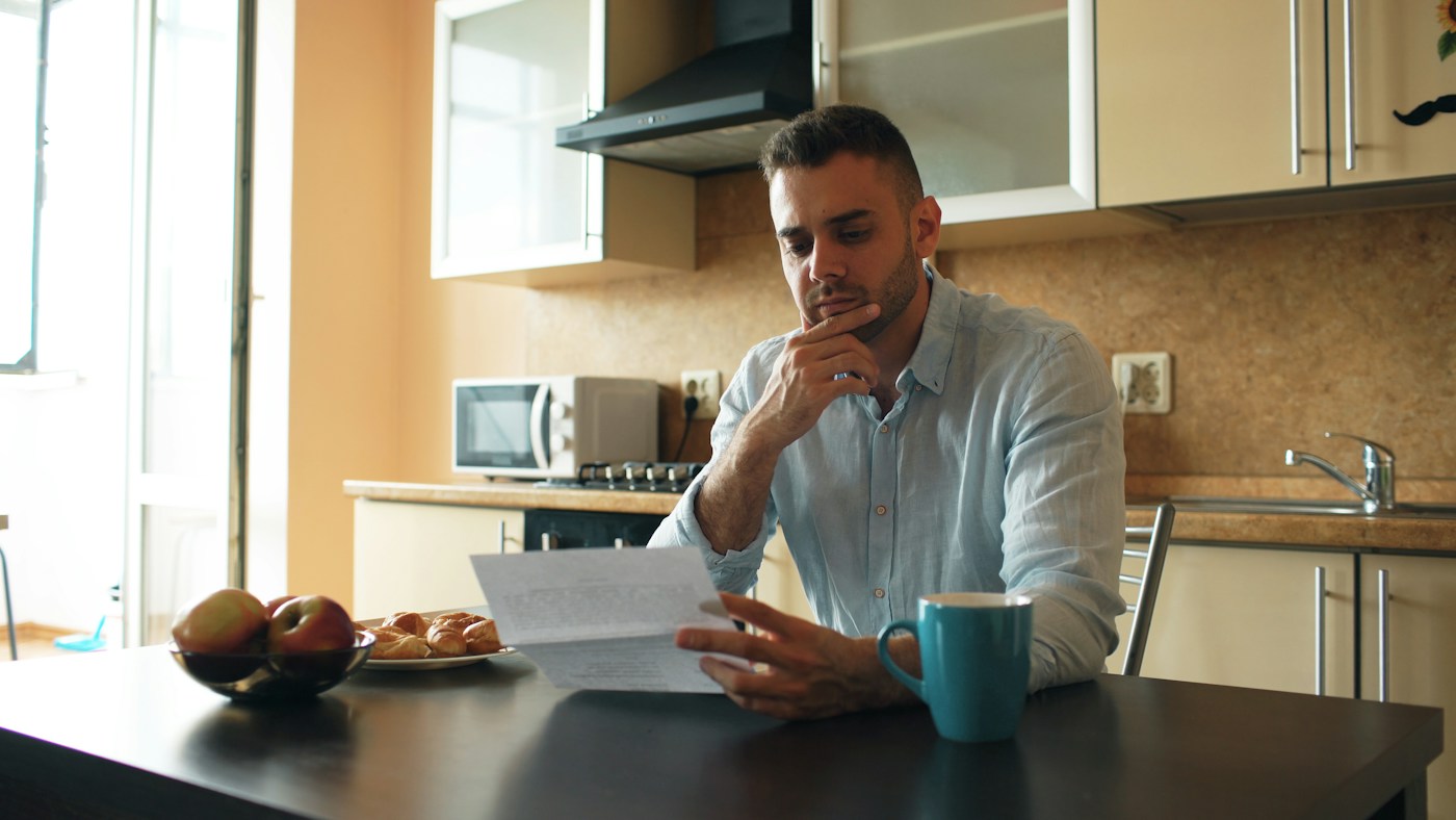 person reading a printed document at a kitchen table with a calculator