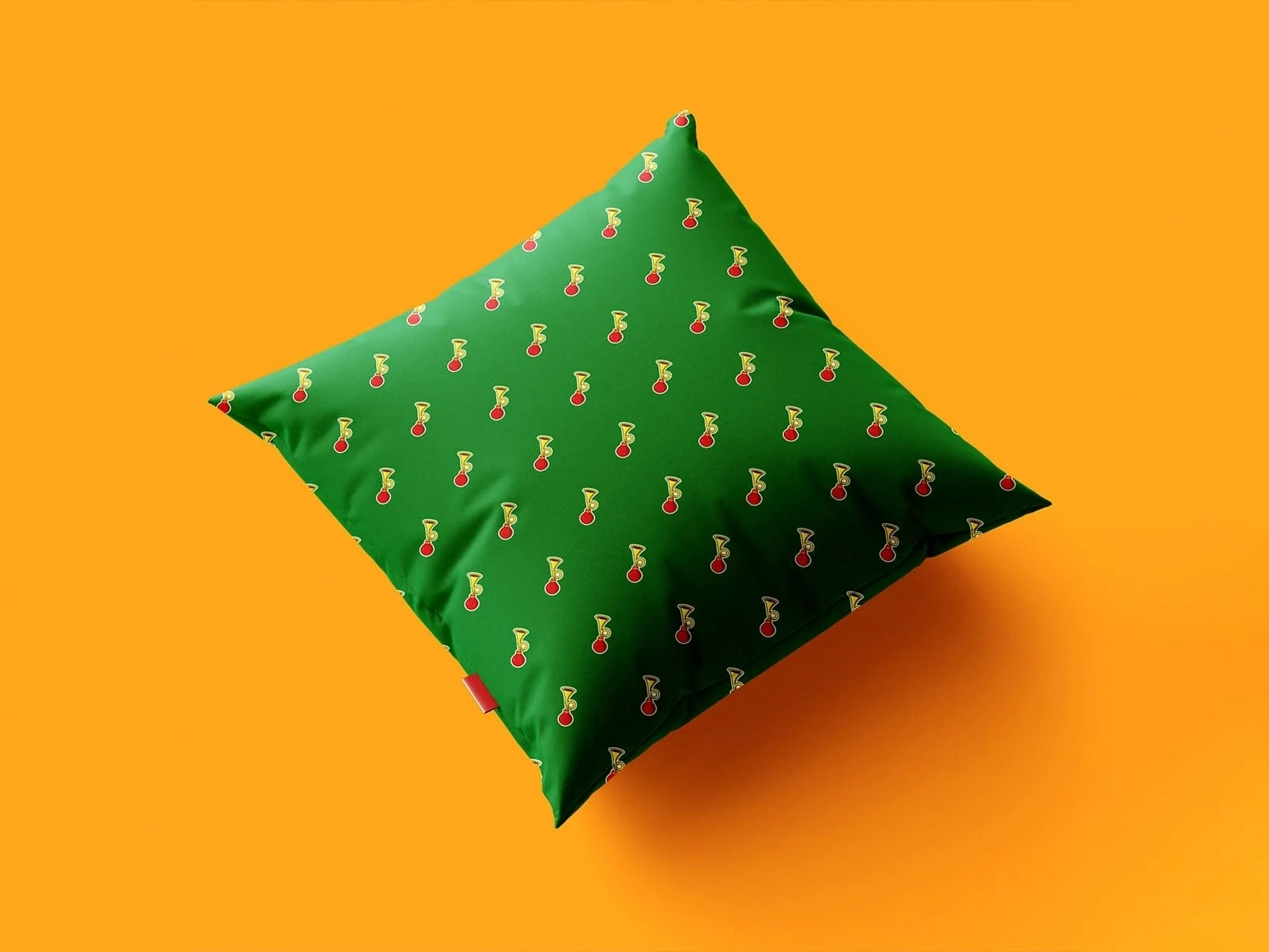 A green square cushion with tiny red-and-white festive motifs floats above an orange surface, casting a soft shadow in a warm, playful scene.