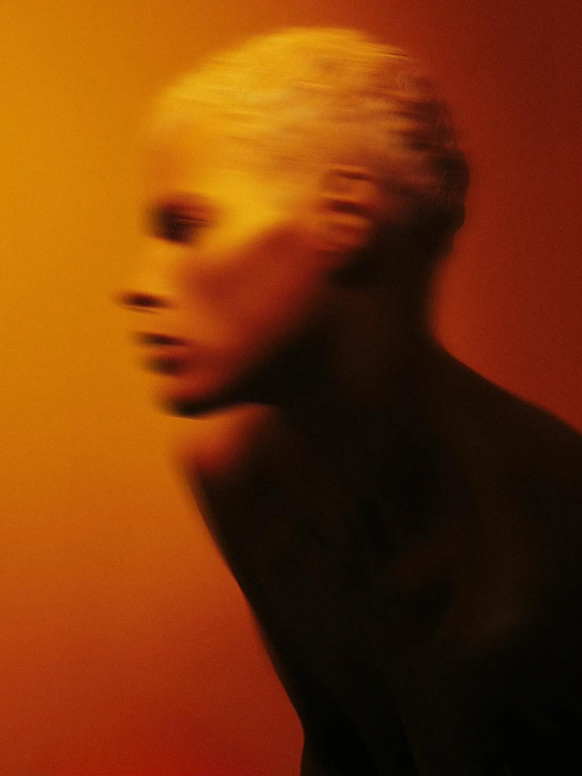 Motion-blurred portrait with short blonde hair against an amber background.