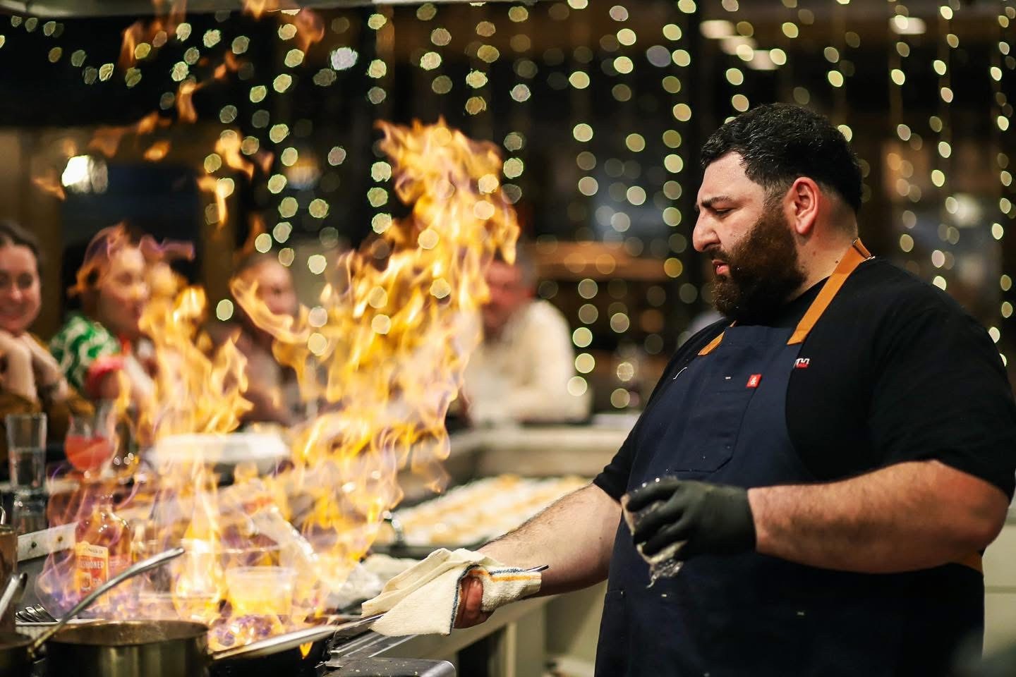 A professional chef with a beard wearing a black apron and gloves, focused on a large flambé fire rising from a pan on a restaurant stovetop while diners watch in the background.