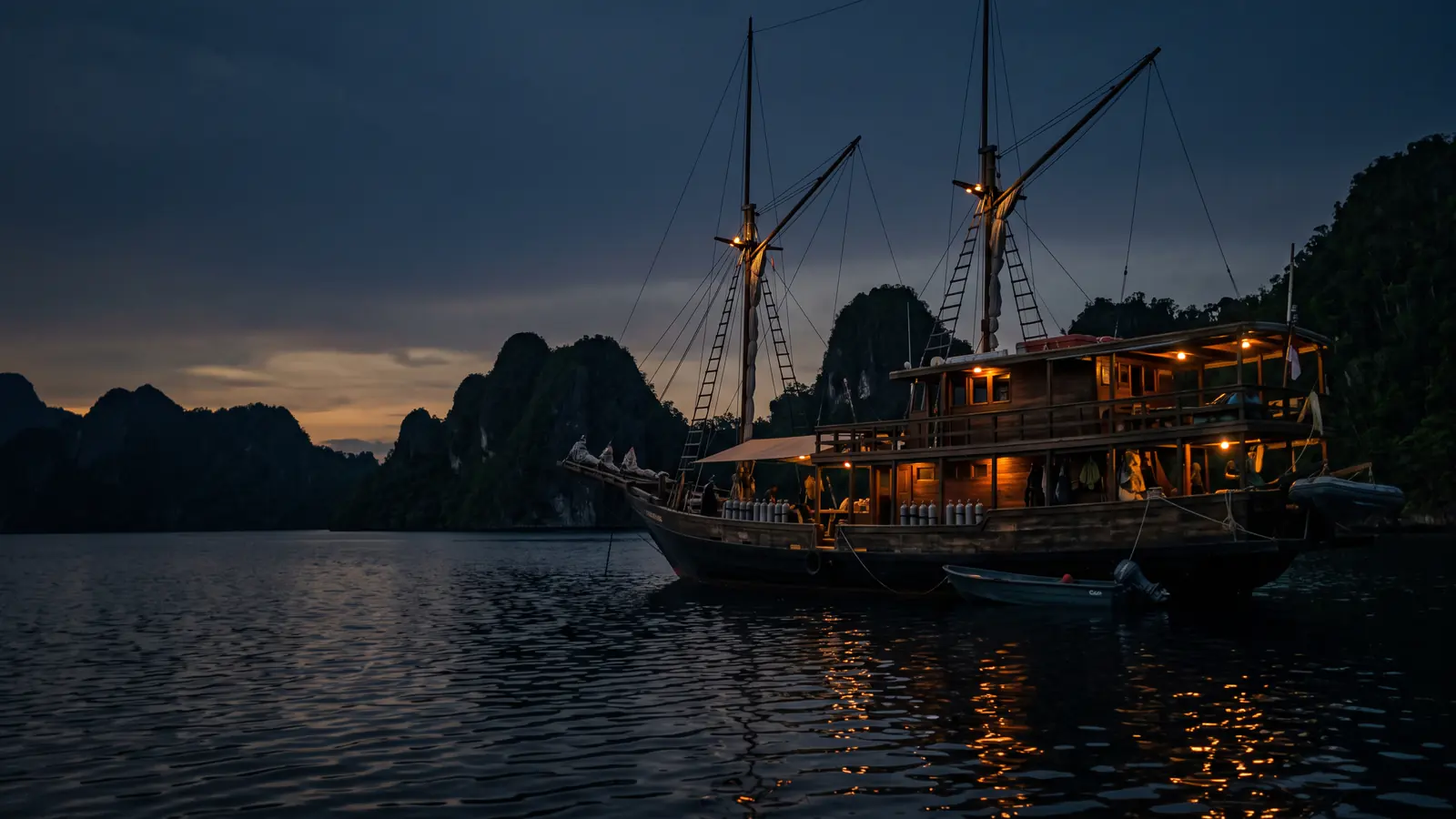 A wooden phinisi at anchor with its lamps lit at dusk