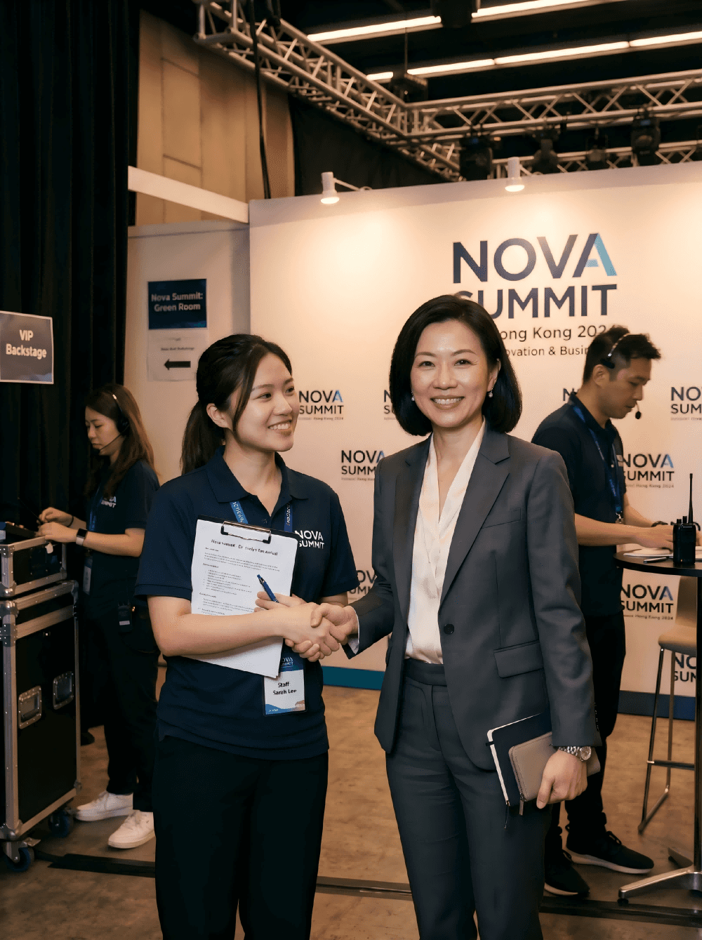 香港商務活動重磅嘉賓接待場景，一位亞洲女性主講嘉賓在後台以得體笑容與活動代表握手，展現專業VIP接待與真實商務交流氛圍 Asian female keynote speaker backstage at Hong Kong corporate event with professional VIP reception and authentic business hospitality