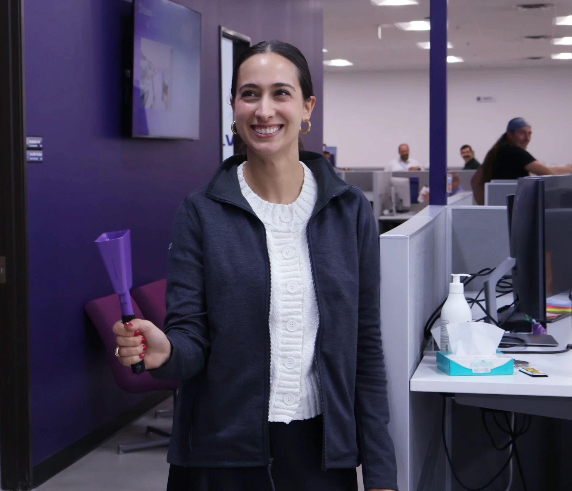 A smiling woman holds a purple bell in an office, with coworkers at their desks behind her.