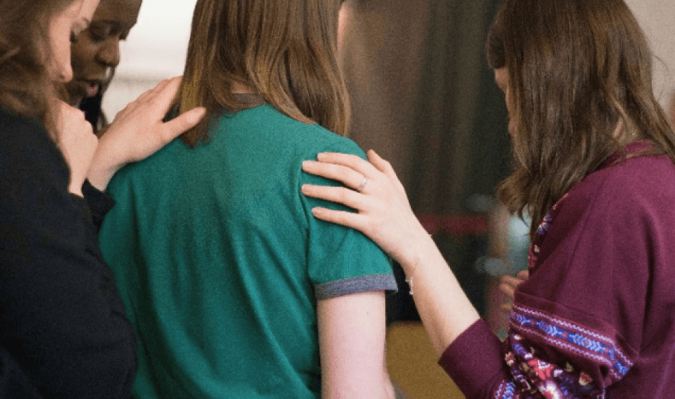 A girl in a green shirt surrounded by three women offering comfort, hands gently on her shoulders.