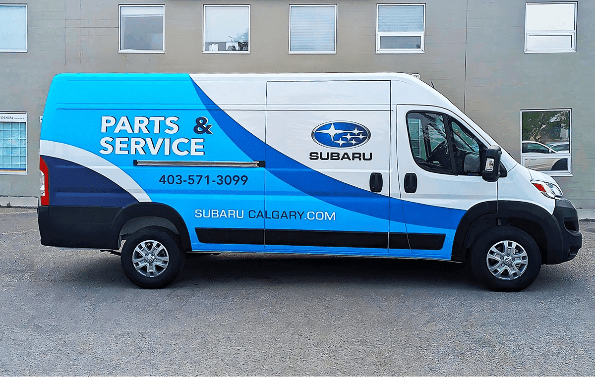 A Dodge windowless white van with white and blue vinyl graphics for Subaru, side view