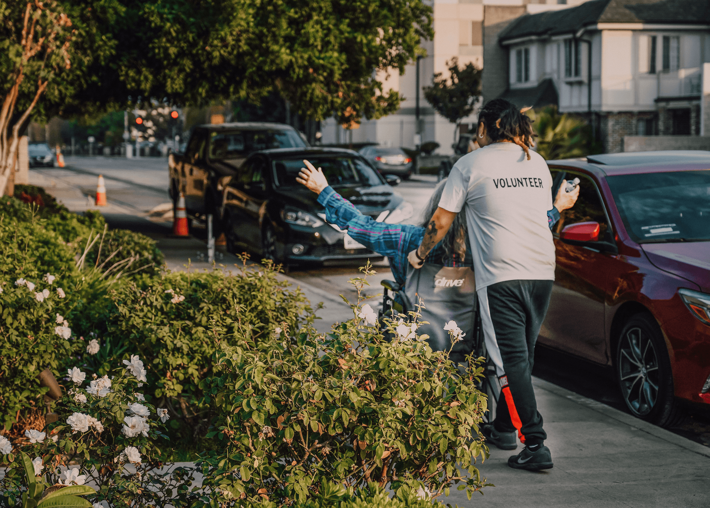 A volunteer assisting a person in a mobility chair along a city sidewalk