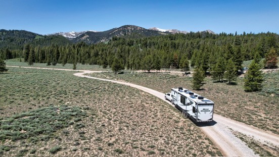 An RV on a winding dirt road through an open meadow beside evergreen forest and mountains.
