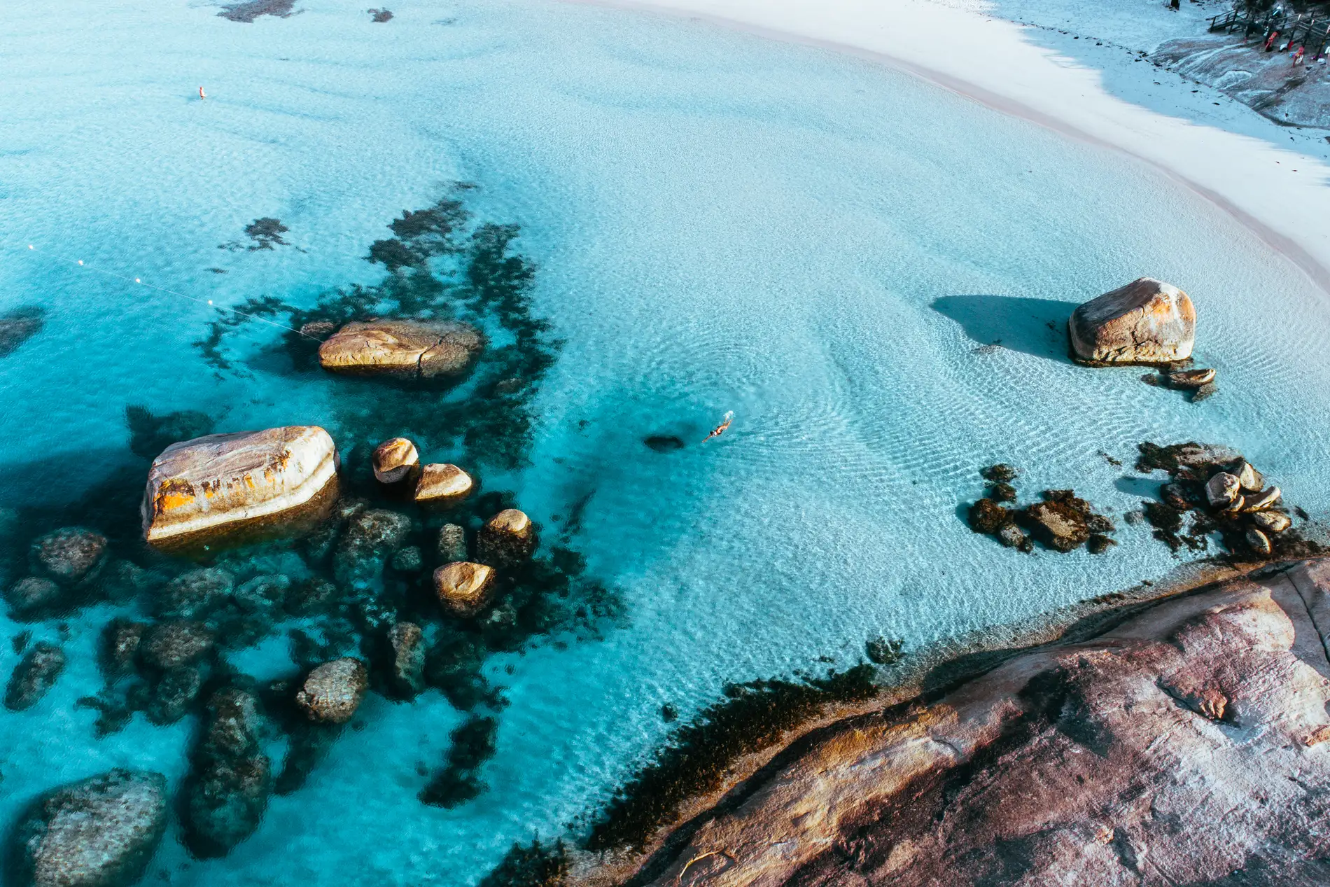 Aerial of granite boulders scattered through shallow turquoise water beside a sandy shore