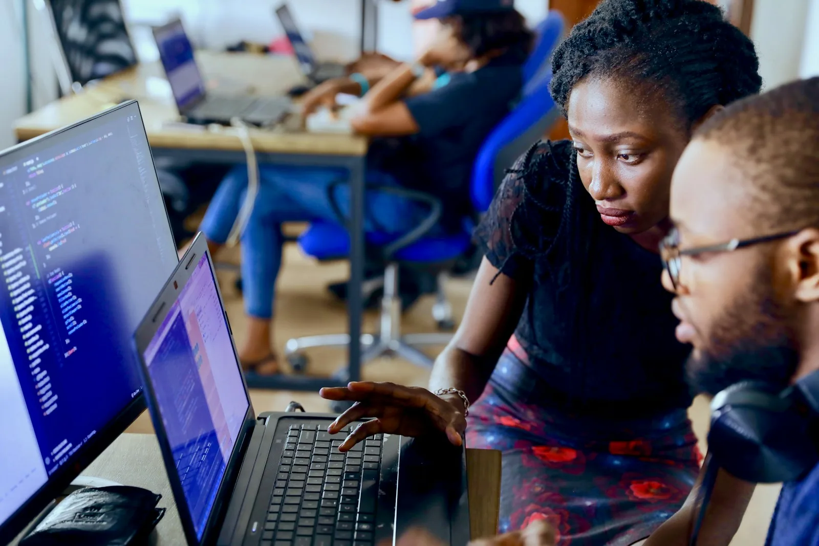 Two colleagues reviewing work together on a laptop and a monitor in a shared studio, with a teammate working in the background