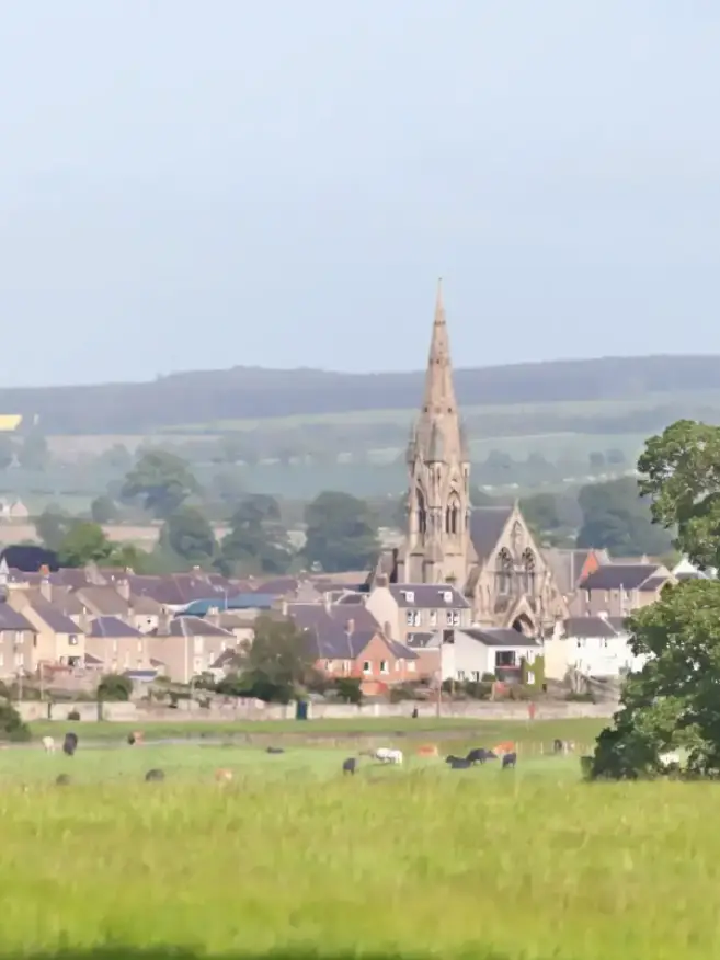 Scenic view of a rural village with a prominent church spire rising above houses. Lush green fields and distant rolling hills create a serene atmosphere.