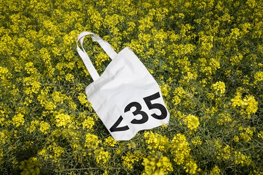 White tote bag with Less Than 35 bold typography among yellow flowers — cultural branding by OUF Design Studio