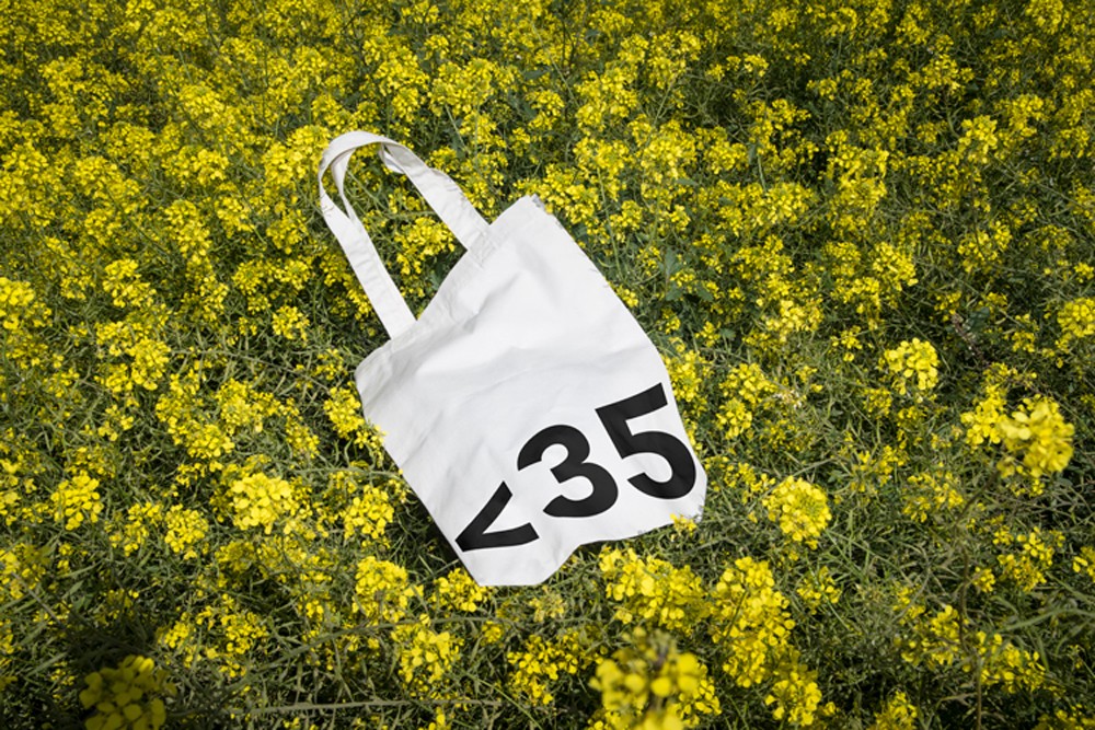 A white tote bag with the Less Than 35 logo and bold black typography lying among bright yellow flowers in an outdoor field setting.