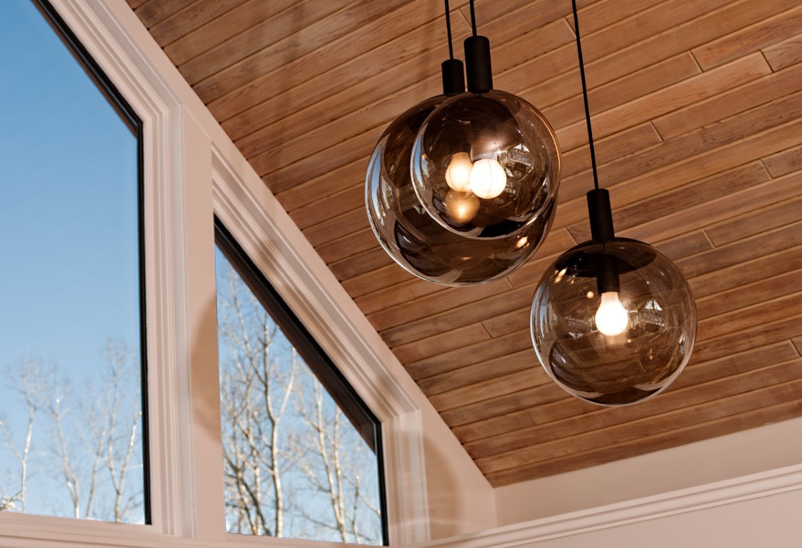 Image of Sleek glass pendant lights hang from wooden ceiling with large windows overlooking trees outside KHD Homes design.