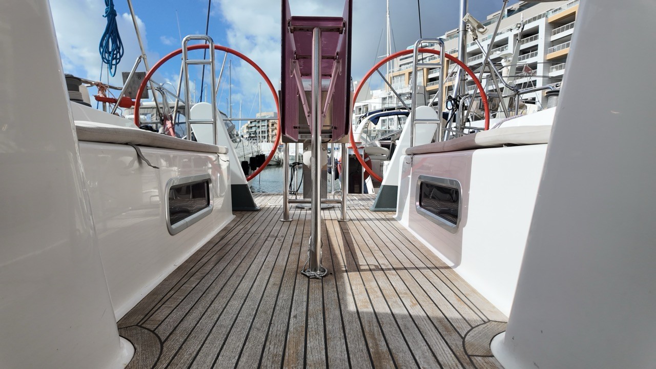 Aft deck of the Hanse 470e looking forward between twin helm stations fitted with red steering wheels, showing EcoDeck teak-tone composite deck planking running the full width of the cockpit sole, with portlight windows in the coamings