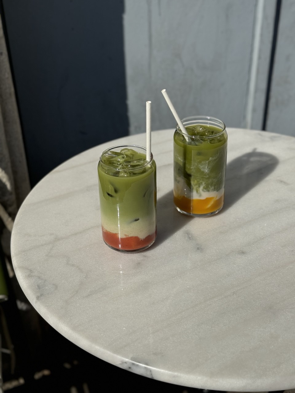 Two glasses of iced matcha drinks with layers of red and orange fruit puree sit on a round white marble table in natural sunlight, casting soft shadows.