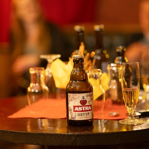 Bottle of Astra beer on a table with various glasses and bottles in a dimly lit setting; blurred figure in the background.