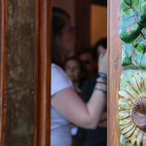 Primer plano de un marco de puerta tallado con un detalle de girasol. A través de la entrada, se vislumbra a una persona desenfocada con una camisa blanca y gafas.