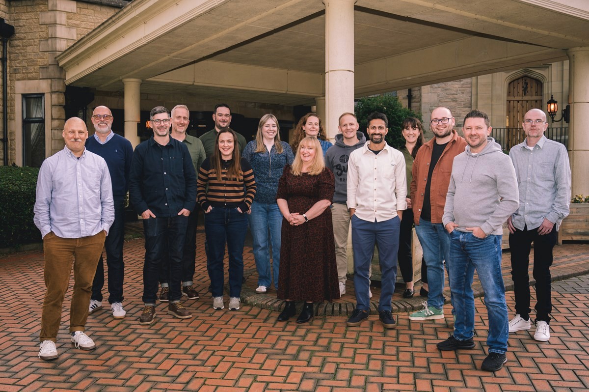 Opencast leadership group standing together outdoors on a brick‑paved area in front of a covered entrance to a stately building. The group is arranged in a loose line, dressed in smart‑casual clothing including shirts, jumpers, jeans, and coats. Stone columns and a wooden doorway frame the background, suggesting a formal venue such as a hotel or conference location. The scene has the feel of a team or group photo taken during an organised off‑site or work event.