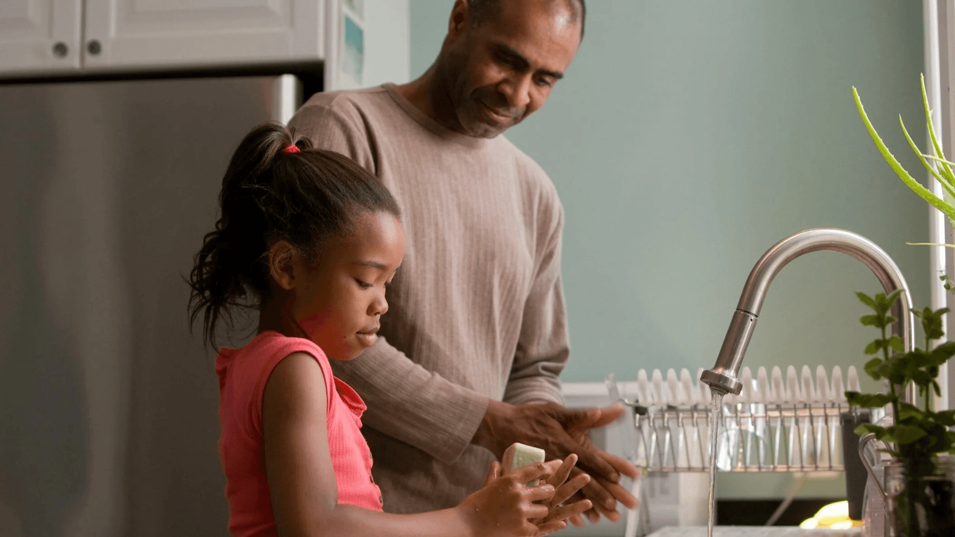 Father and daughter washing their hands together at a kitchen sink, with the child holding a bar of soap while water runs from the faucet.