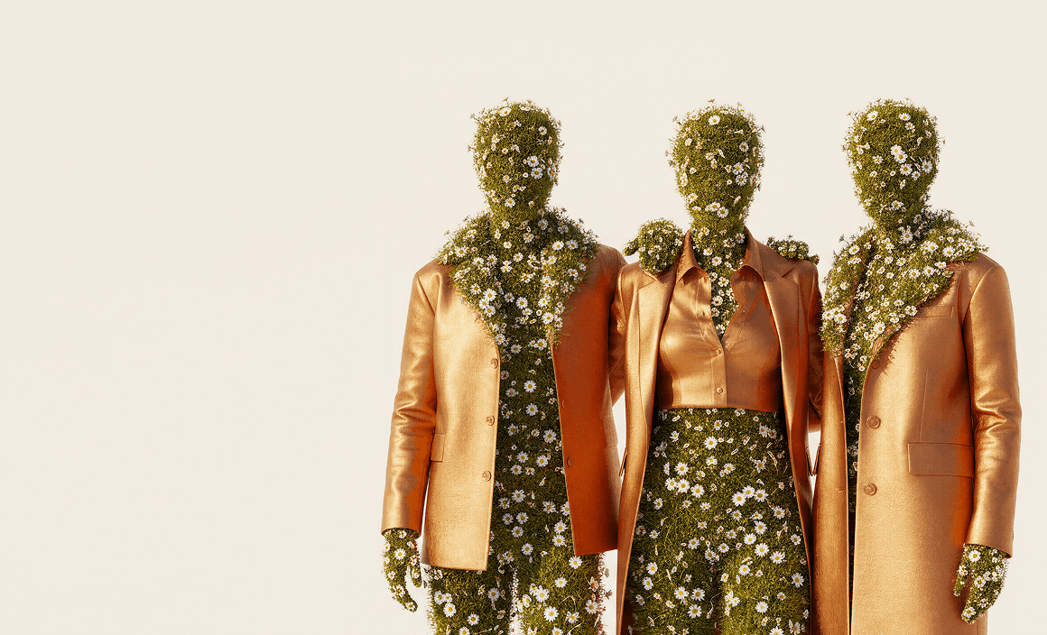 A surrealist digital artwork of three humanoid figures standing side-by-side against a cream background. Each figure is composed of green grass and blooming white daisies where skin would be, including their heads and hands. They are dressed in fashionable, metallic bronze-orange suits and a crop-top blouse. The figure in the center has their arms around the shoulders of the other two, and daisy vines spill over the lapels and collars of their jackets.