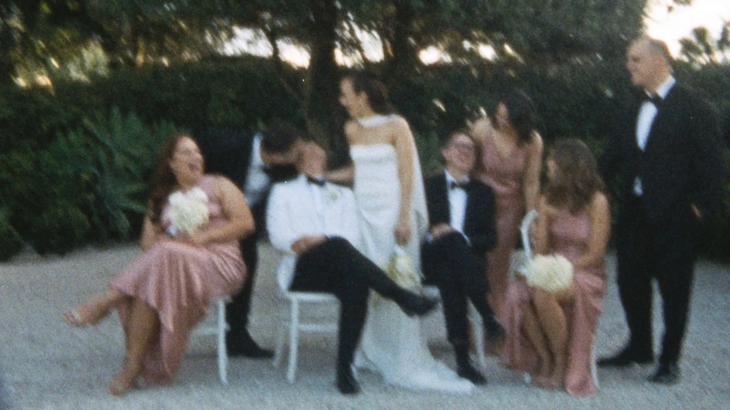 A group of people dressed elegantly for a wedding gather outdoors, with women in pink dresses holding bouquets and men in dark suits and tuxedos, while trees and greenery provide a lush backdrop.