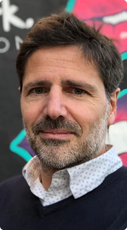 A close-up portrait of a middle-aged man with dark, salt-and-pepper hair and a groomed beard, wearing a white patterned shirt under a dark blue v-neck sweater.