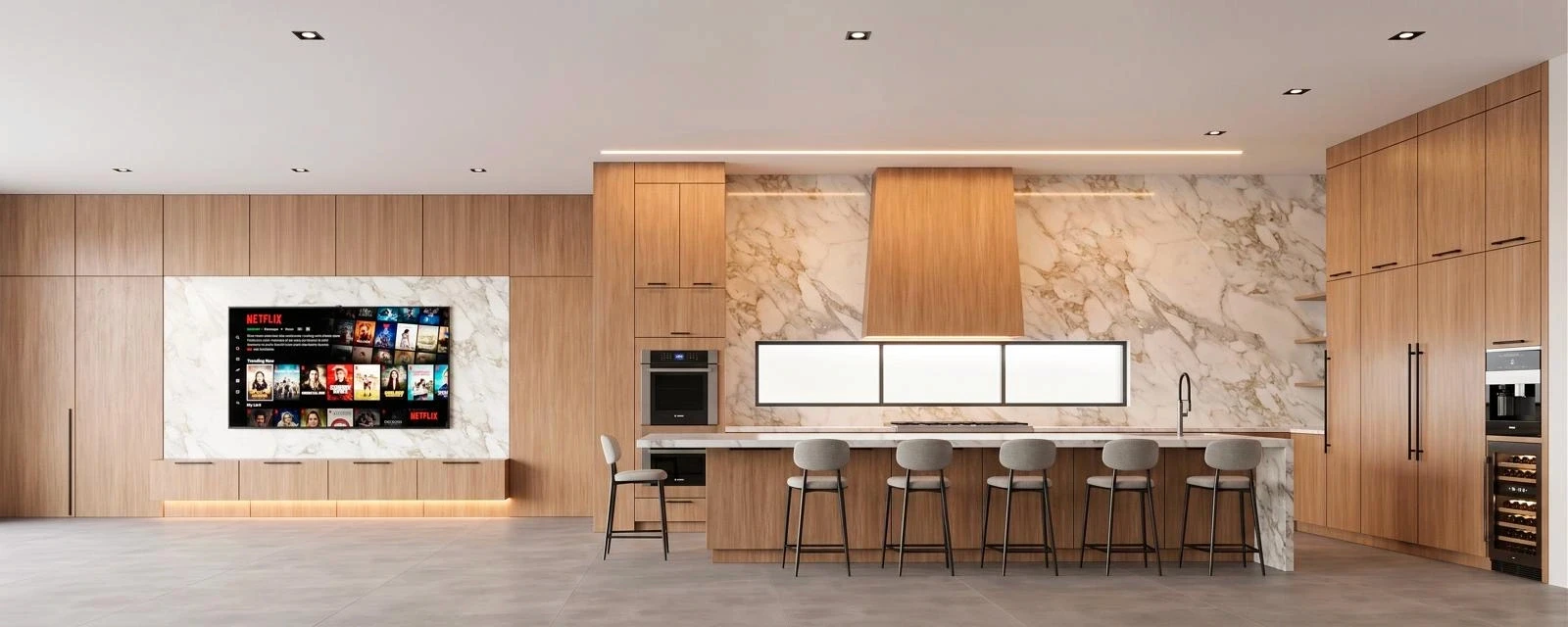 Modern open-plan kitchen with light wood cabinetry, a large marble-backed island with five upholstered bar stools, a wood-panel range hood, and a wall-mounted TV set into a marble recess displaying a streaming interface.