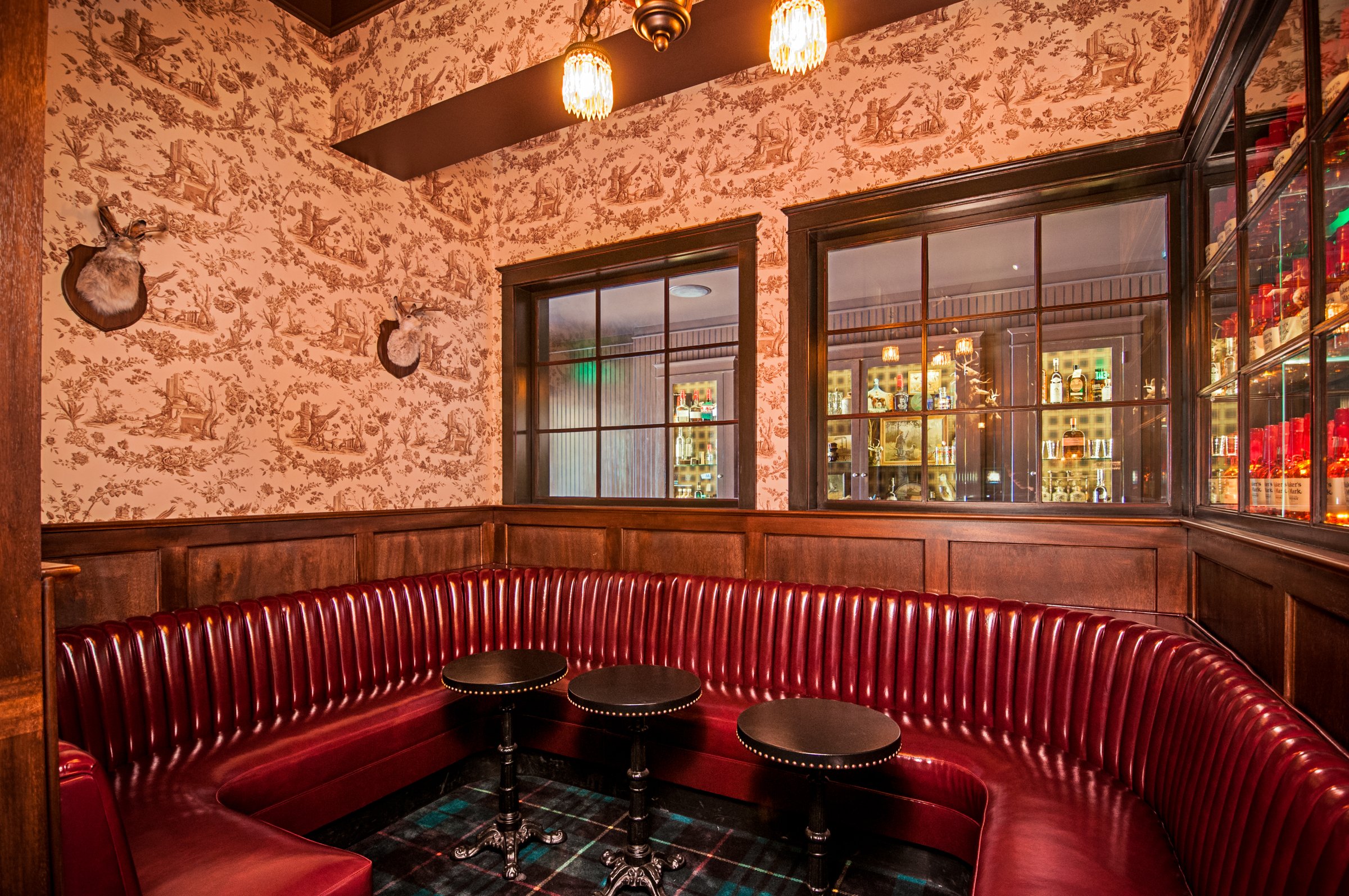 Curved red leather banquette with small round tables, dark timber paneling and patterned wallpaper.