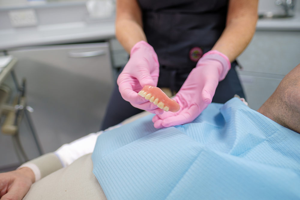 A dental professional wearing pink gloves holds a set of dentures over a patient covered with a light blue drape in a modern dental office.