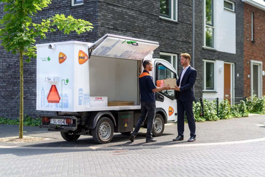 PostNL employee delivers a parcel to a man wearing a suit in front of an open PostNL truck in a residential area