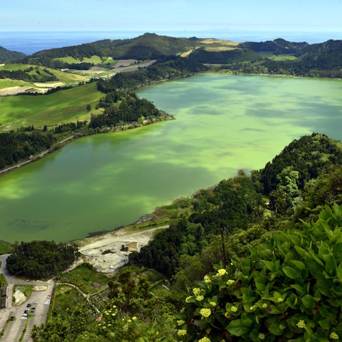 بحيرة زاهية خضراء محاطة بتلال مورقة ونباتات، مع طريق صغير ومباني في الزاوية السفلية اليسرى.