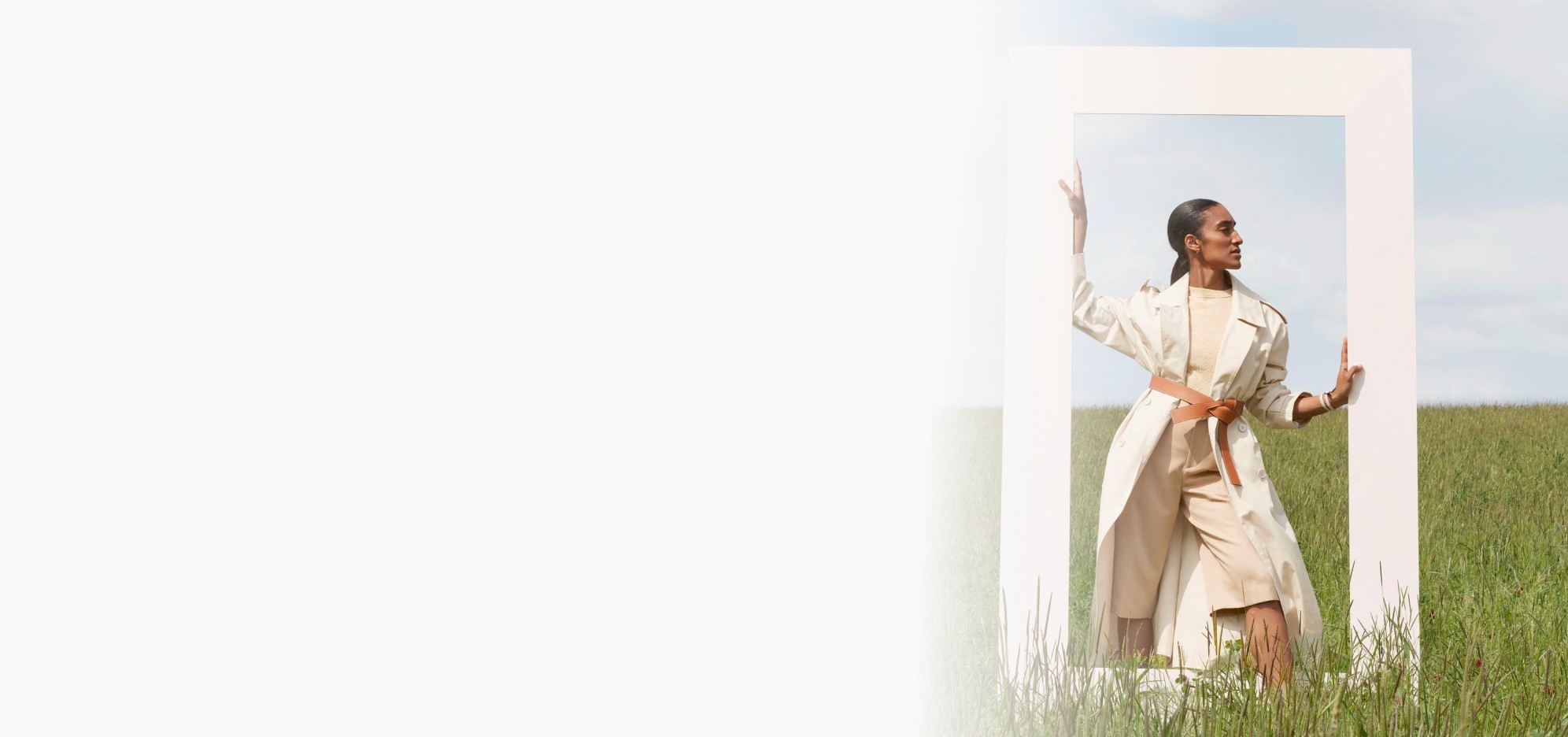 Femme élégante en trench beige posant dans un cadre blanc au milieu d’un champ