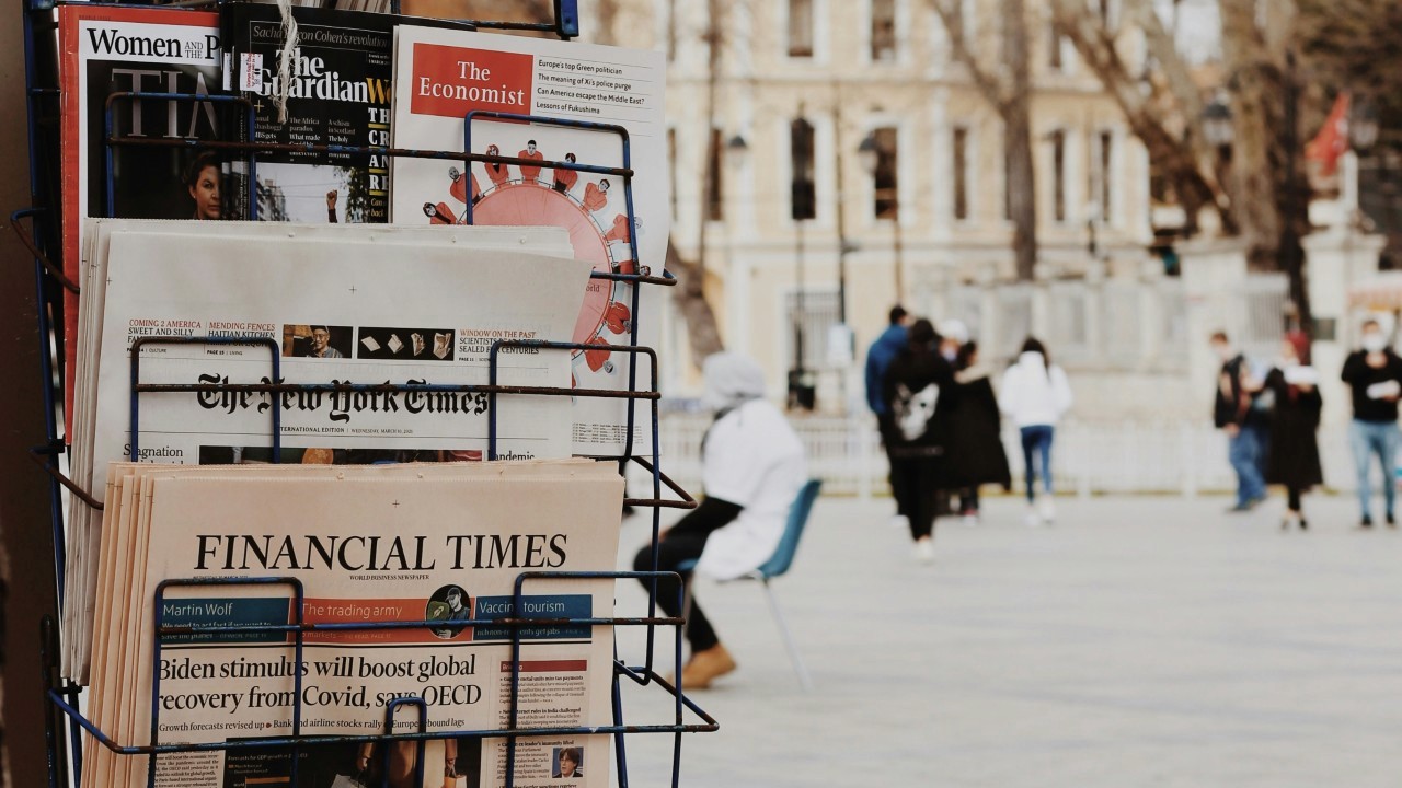Close-up of a newsstand selling international titles, with people passing by on the streets in the background