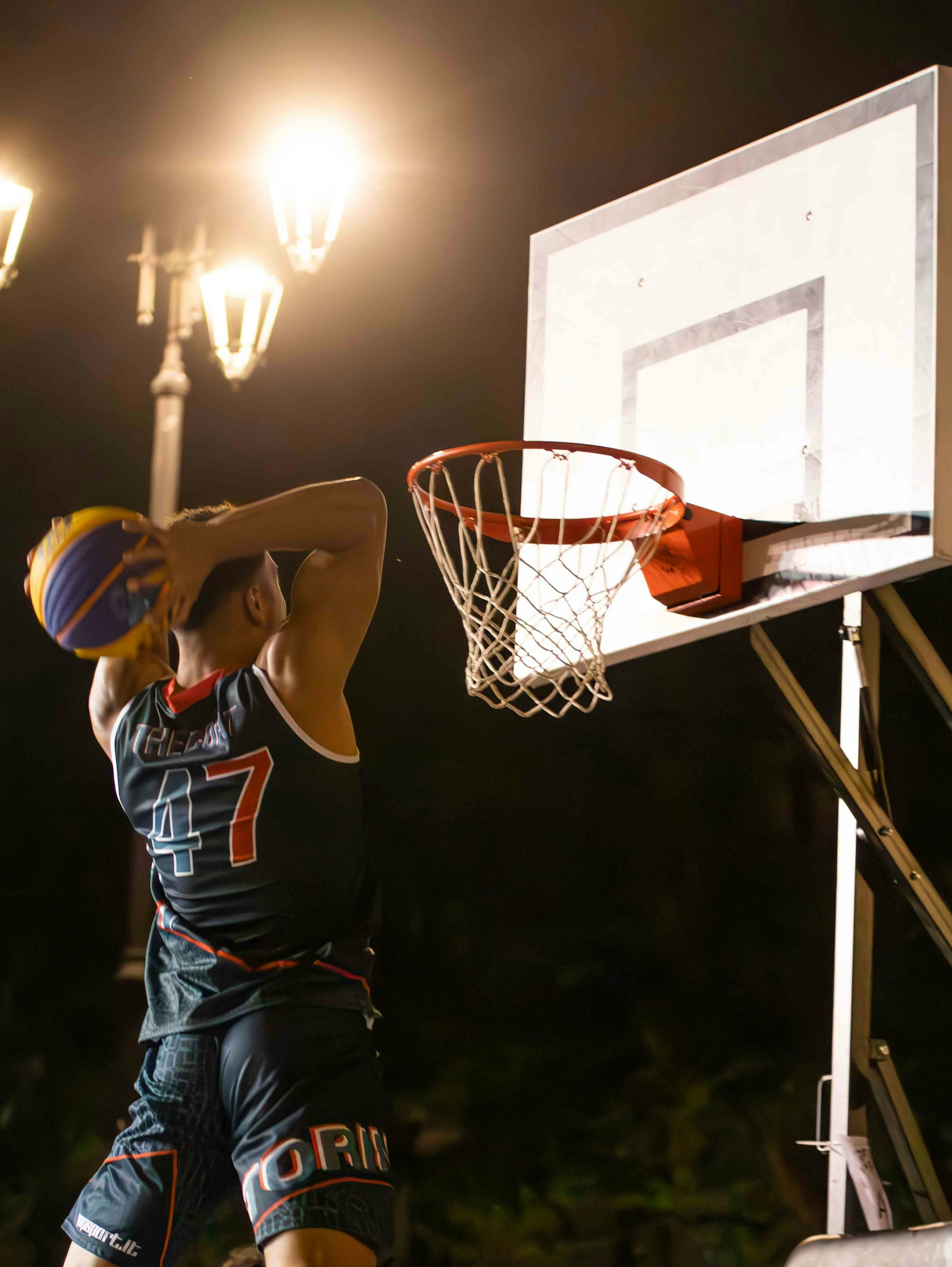 Rear view of a basketball player in a dark #47 jersey dunking on an outdoor hoop at night under streetlights.
