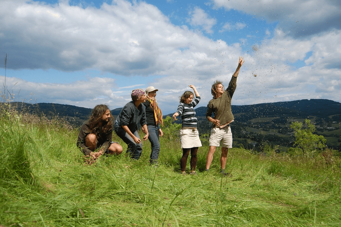 A young person with curly hair smiles at the camera while sitting in front of a laptop against a dark background.