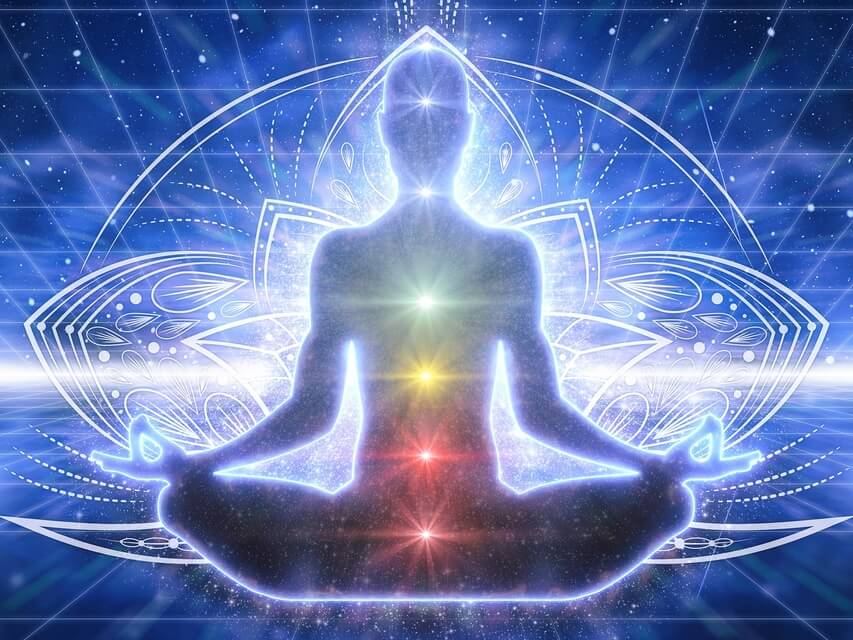 A circle of people sitting cross-legged, meditating with hands in a prayer position. Soft, warm lighting.