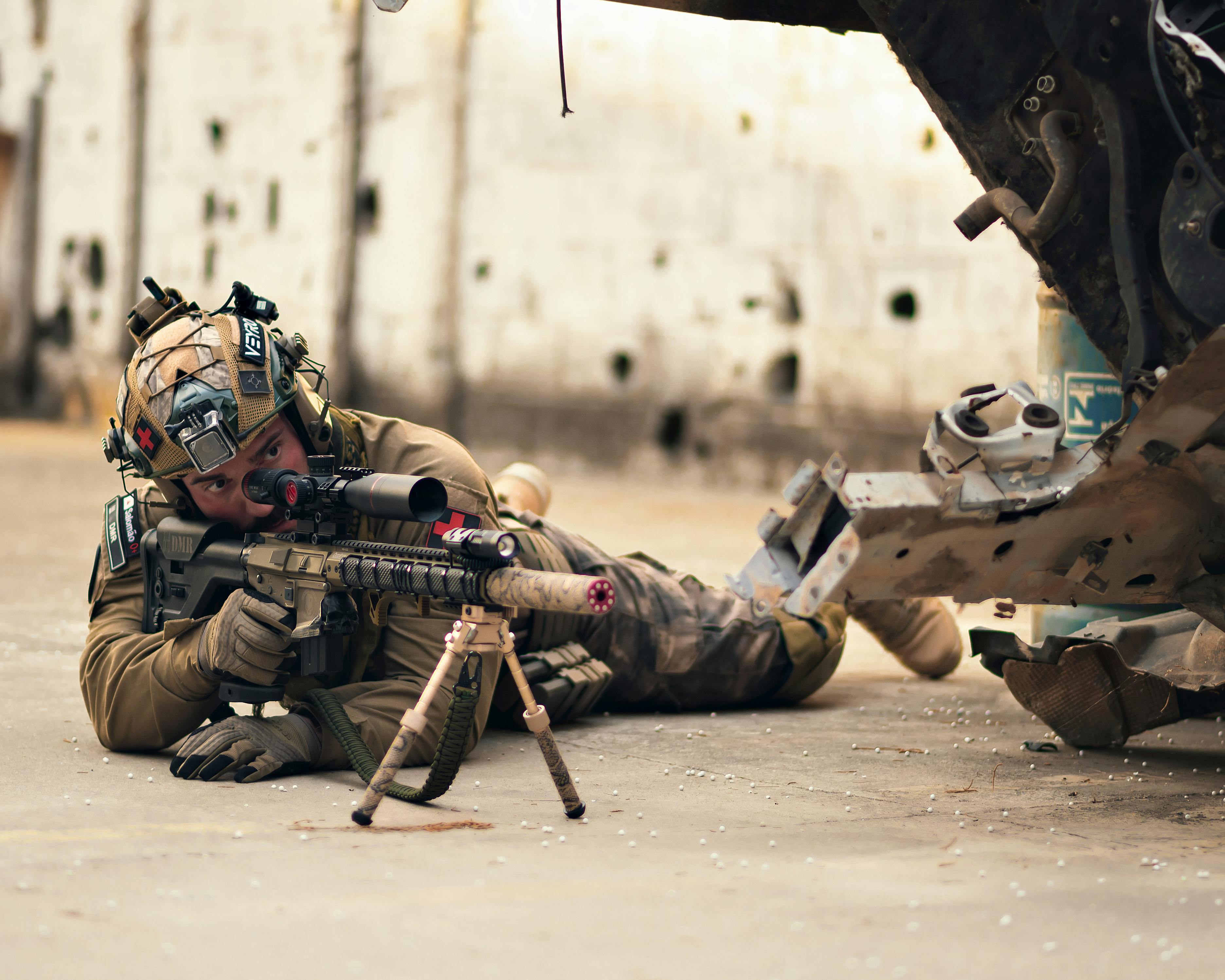 The image shows a military or tactical personnel in a prone firing position on a concrete floor. The person is wearing desert-colored tactical gear including a combat helmet with various attachments. They are aiming a rifle equipped with a scope and bipod support. The rifle appears to be configured for precision shooting. The scene takes place in what looks like a damaged or abandoned building with a light-colored wall that shows signs of damage or bullet holes in the background. On the right side of the image, there appears to be part of a vehicle or piece of equipment. The overall setting suggests this could be a military training exercise, combat simulation, or possibly actual combat operations in an urban environment.