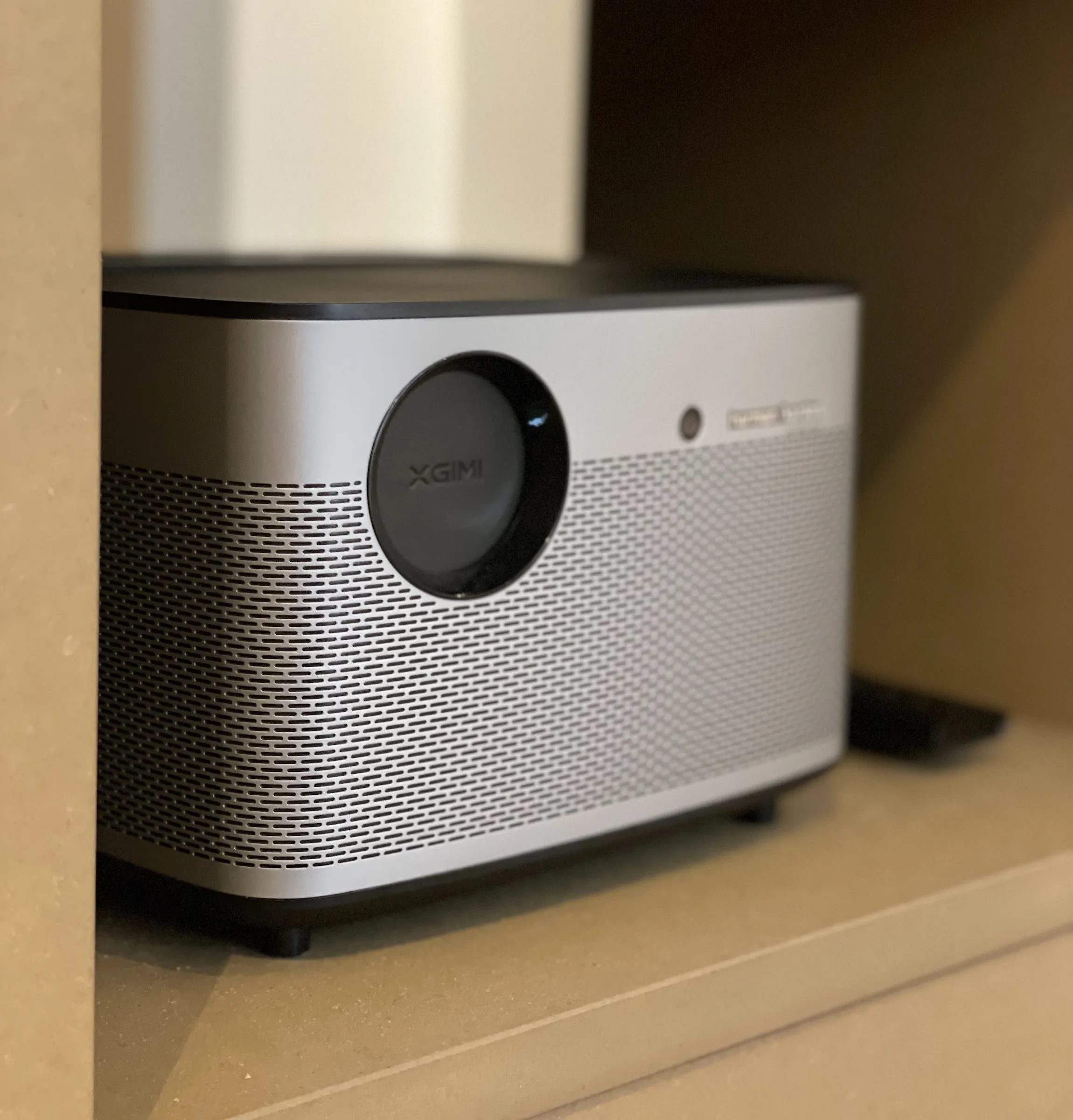 Ein eleganter, silberner XGIMI-Projektor steht auf einem braunen Regal, ausgestattet mit einer runden Linse und einem perforierten Lautsprechergitter. Die Stimmung ist modern und technikorientiert.