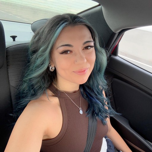 Young person with wavy, shoulder-length red hair, wearing a rust-colored vest over a beige shirt. A brown scarf is tied neatly at the neck. Neutral backdrop.